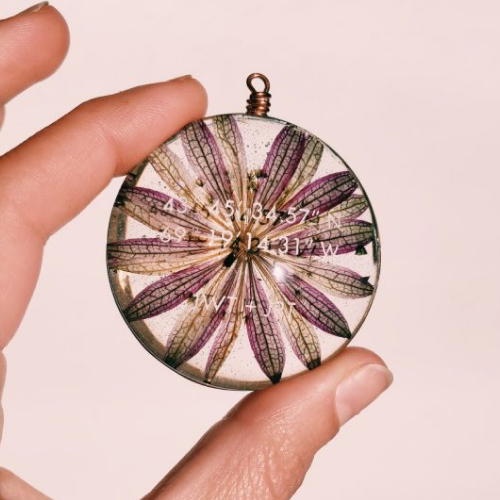 White hand holding a glass pendant with text etched into it and a purple flower in it on a light pink background