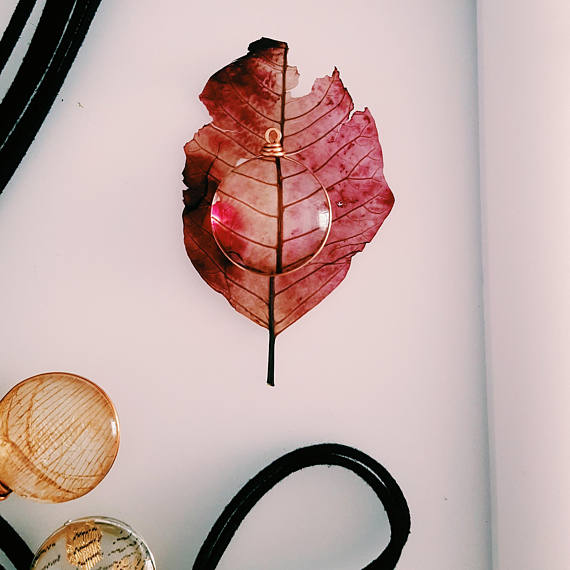 Glass flower pendant laying on a pink leaf on a white background surrounded by another glass petal pendant and leather cords