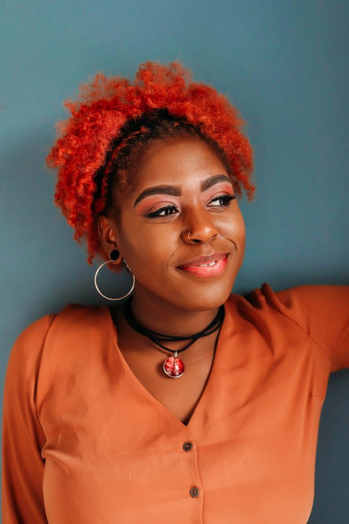 Black girl with red hair wearing an orange blouse and a Gilded Poinsettia Pendant on a black leather chord with a blue background