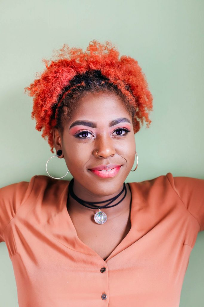 Close up ofBlack girl with red hair wearing an orange shirt and wearing a glitter pendant on a black cord with a light green background
