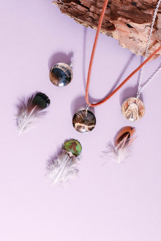 three different colored gilded pheasant feather pendants laying next to feather by their original pheasant feathers on a purple background