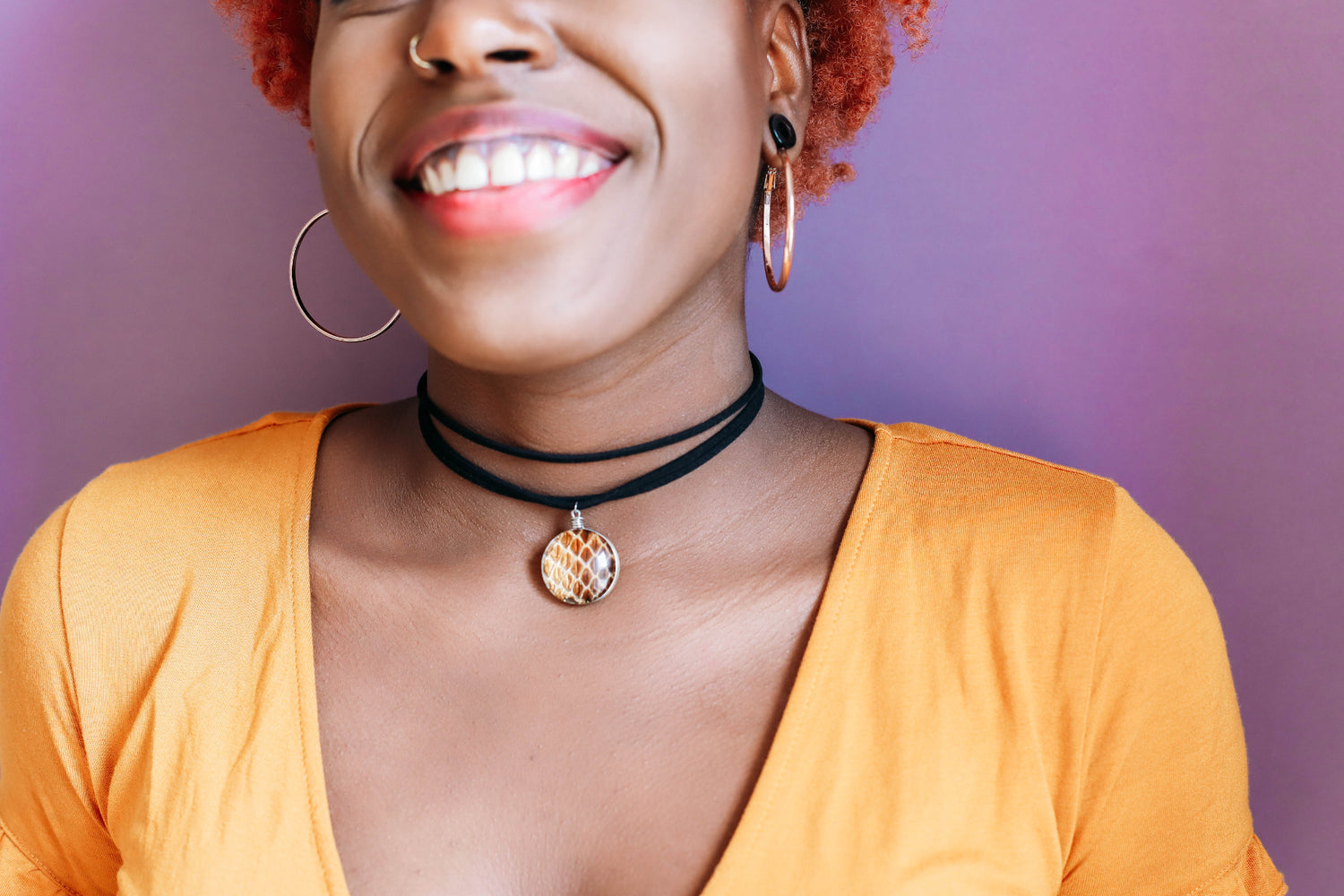 Smiling black girl with red hair wearing a black leather choker necklace with a snake skin glass pendant and orange shirt on a purple background 
