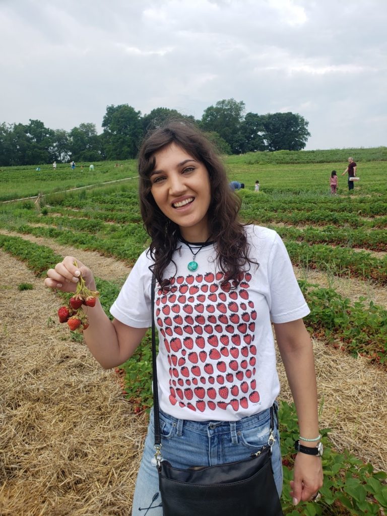 White girl with brown hair in a strawberry field wearing a blue SisTers PGH Rainbow Rose Petal Pendant