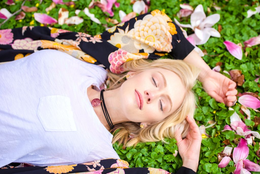 White girl with blond hair wearing a Pink Leaf Pendant on a black cord laying outside on grass and flowers with her eyes closed